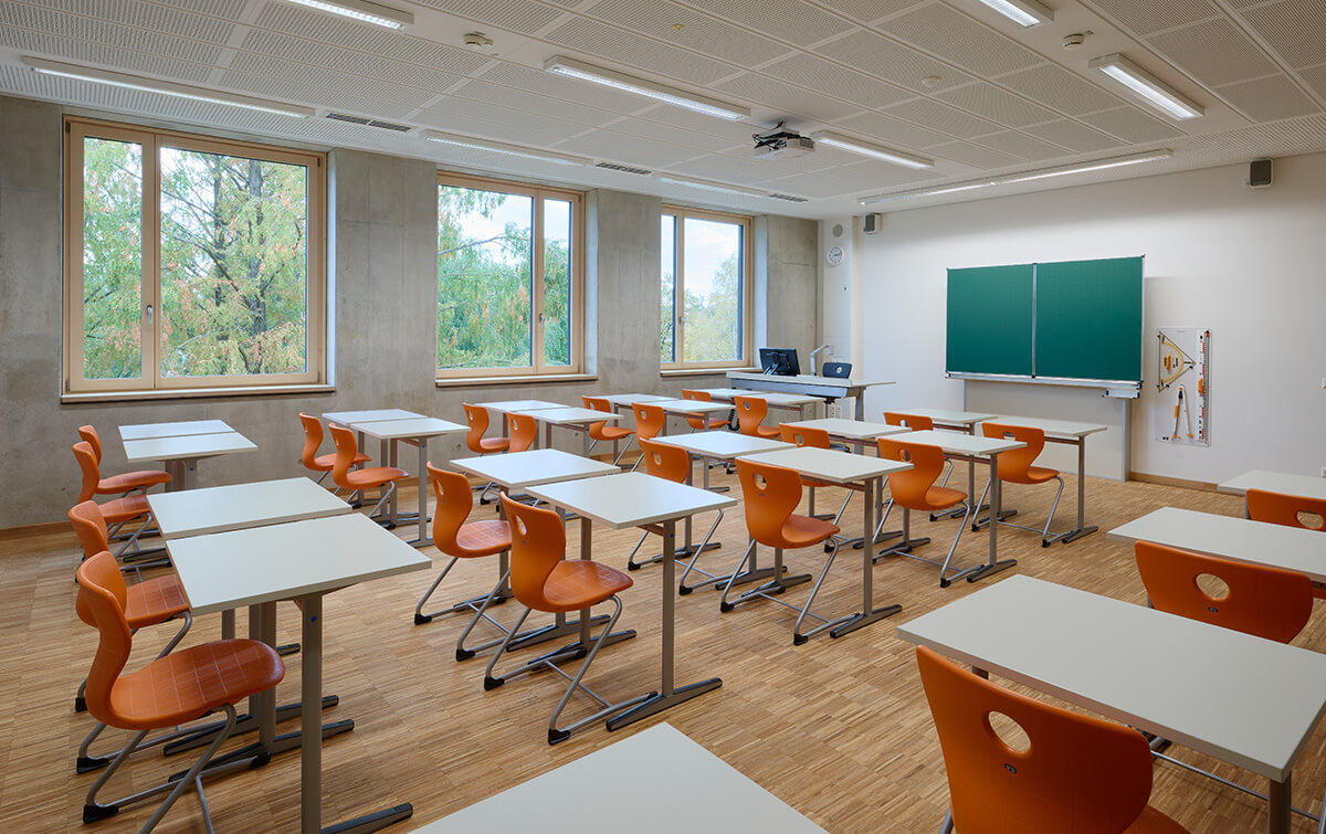 Klassenzimmer der FOS/BOS Krumbach mit Reihen von Tischen und Stühlen, einer grünen Tafel an der Wand und Fenstern, die auf Grünanlagen blicken, beleuchtet durch Deckenleuchten