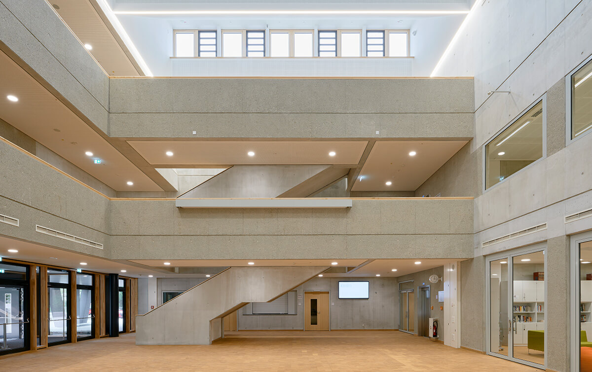 Aula der FOS/BOS Krumbach - Gebäudes mit einer Kombination aus rostroten Metallplatten und dunklen Paneelen, grossen Fenstern und einem Unterbau mit Säulen, vor einem Parkplatz