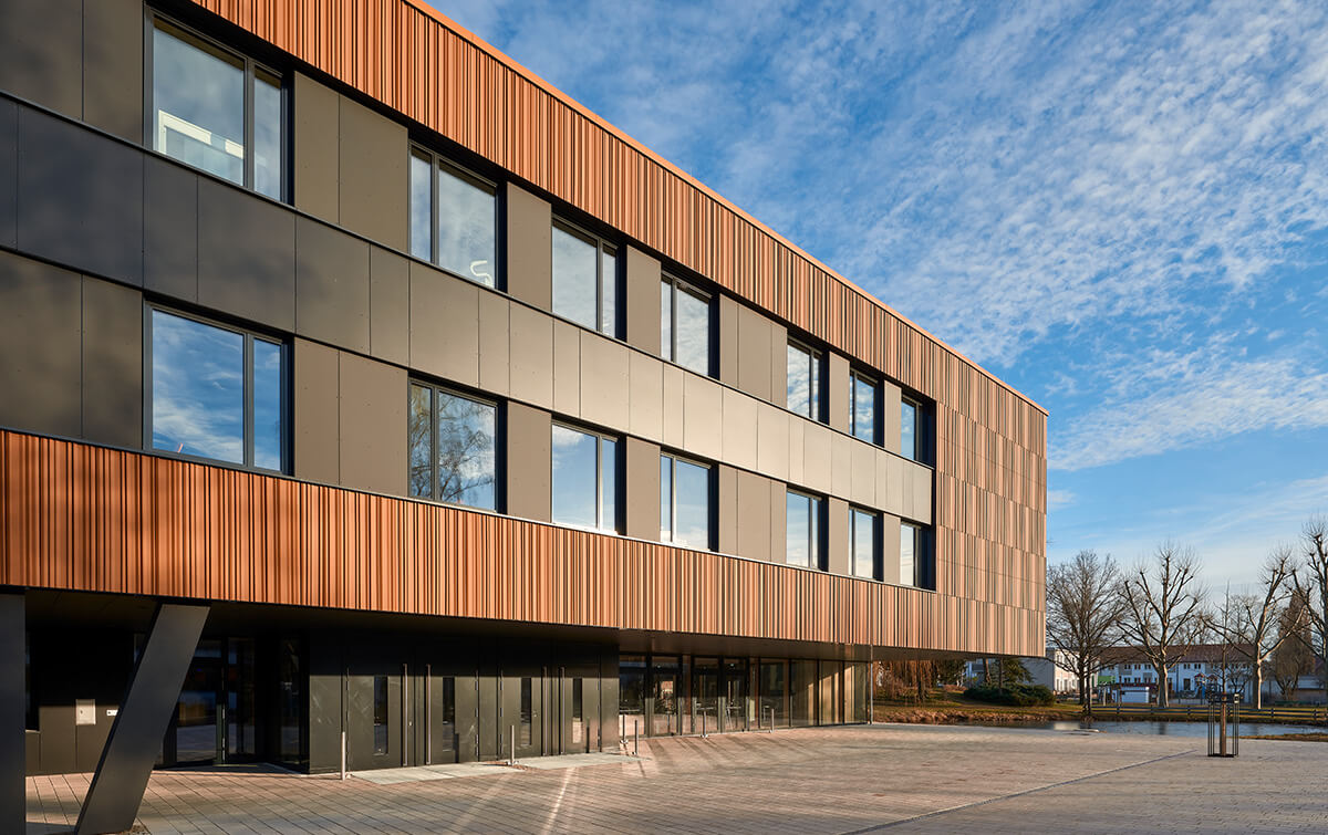 Außenansicht FOS/BOS Krumbach - Gebäudes mit einer Kombination aus rostroten Metallplatten und dunklen Paneelen, grossen Fenstern und einem Unterbau mit Säulen, vor einem Parkplatz