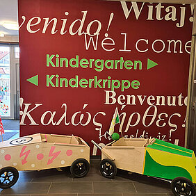 Eingangsbereich eines Kindergartens mit Willkommensschild in verschiedenen Sprachen und zwei bunten Seifenkisten davor