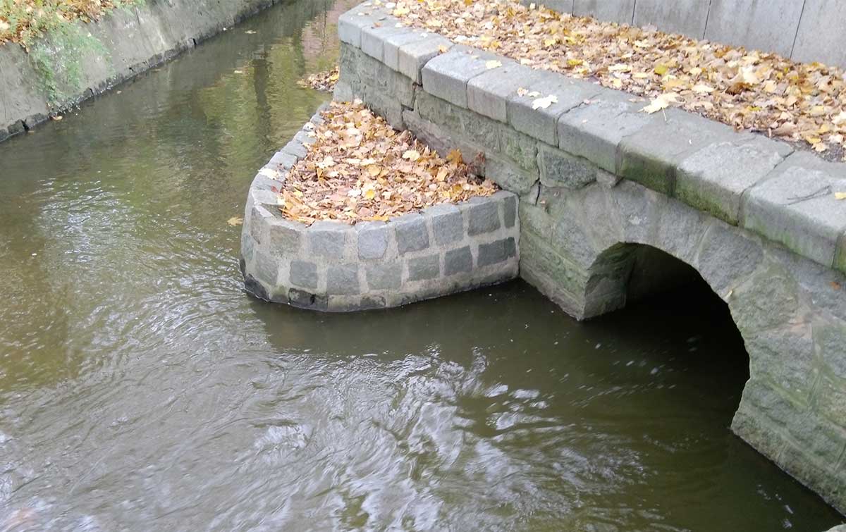 Steinbrücke über einen Fluss, mit einem gemauerten Bogen und Uferbefestigungen aus Stein.